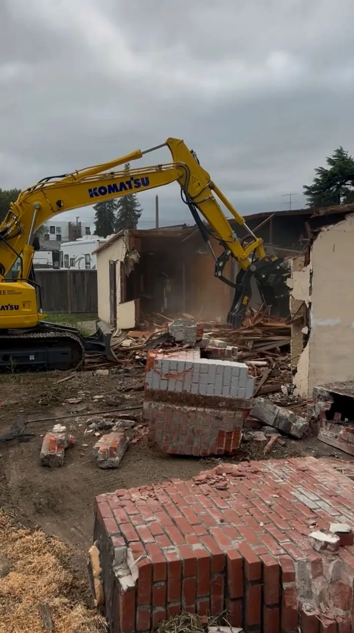 Komatsu excavator pulling down a house, brick chimney rubble in the foreground