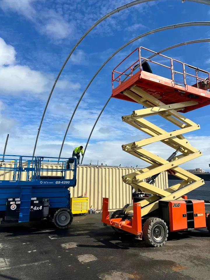 Scissor lifts beside a tent frame, a crew member working on top of a container