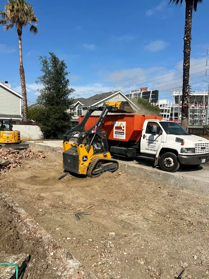 Deere skid steer loading a Mosso roll-off truck on a cleared lot beside Levi's Stadium