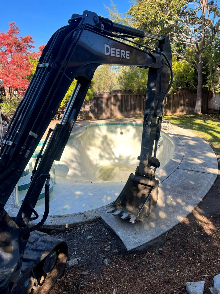 Deere excavator set up at the edge of an empty in-ground swimming pool